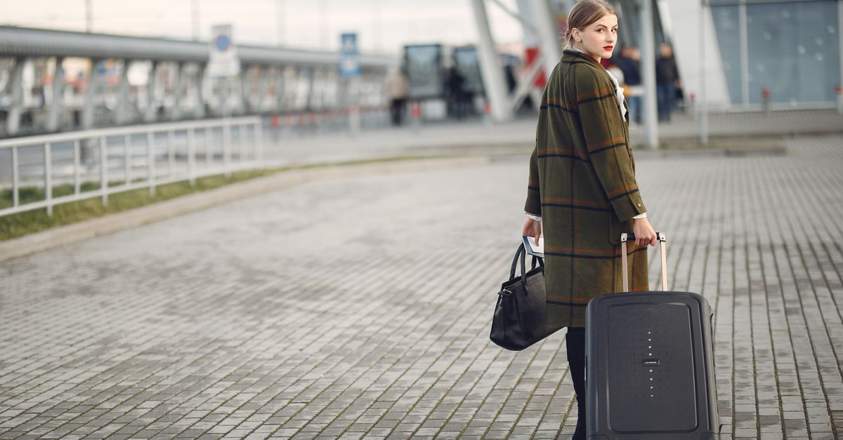 Full length of stylish female traveler in warm coat walking with suitcase and bag looking at camera after arrival against airport terminal