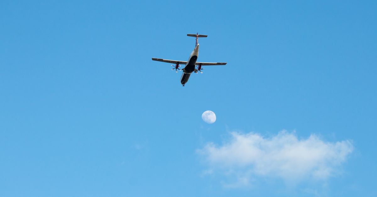 A small airplane soaring in a clear blue sky with the moon visible, captured in London, UK.