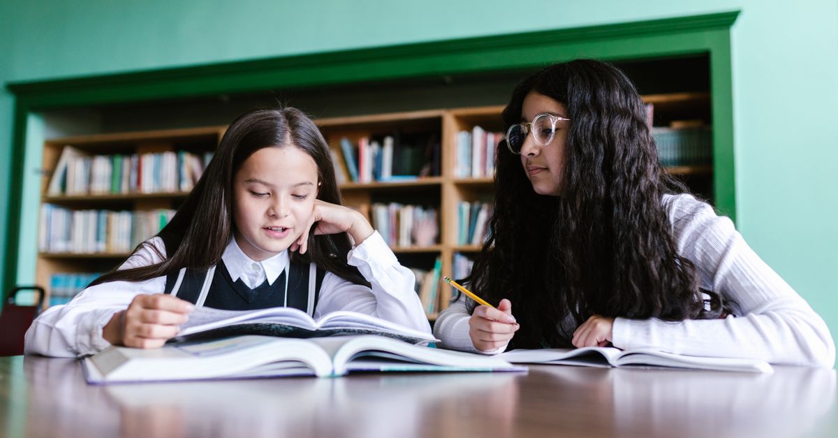 Two girls in school uniforms studying together at a library desk, focused and engaged.