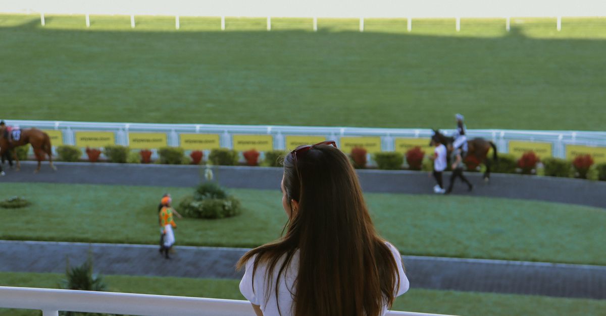 A woman gazes at a horse racetrack, observing jockeys and horses preparing for a race.