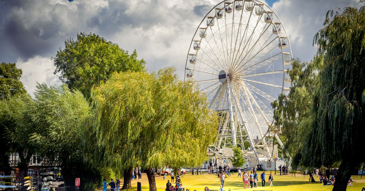 Captured in Stratford-upon-Avon, the Ferris wheel stands tall against a cloudy sky, surrounded by vibrant greenery and lively park-goers.