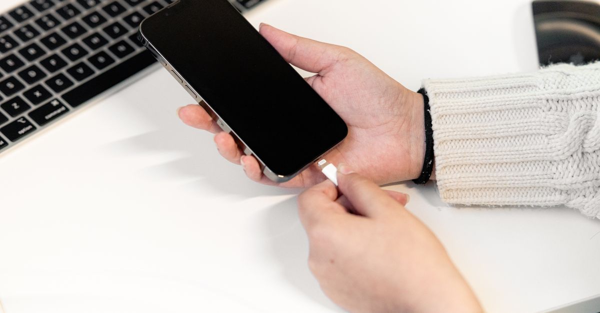 A woman plugs her smartphone into a charger at a desk, next to a laptop, illustrating modern technology usage.